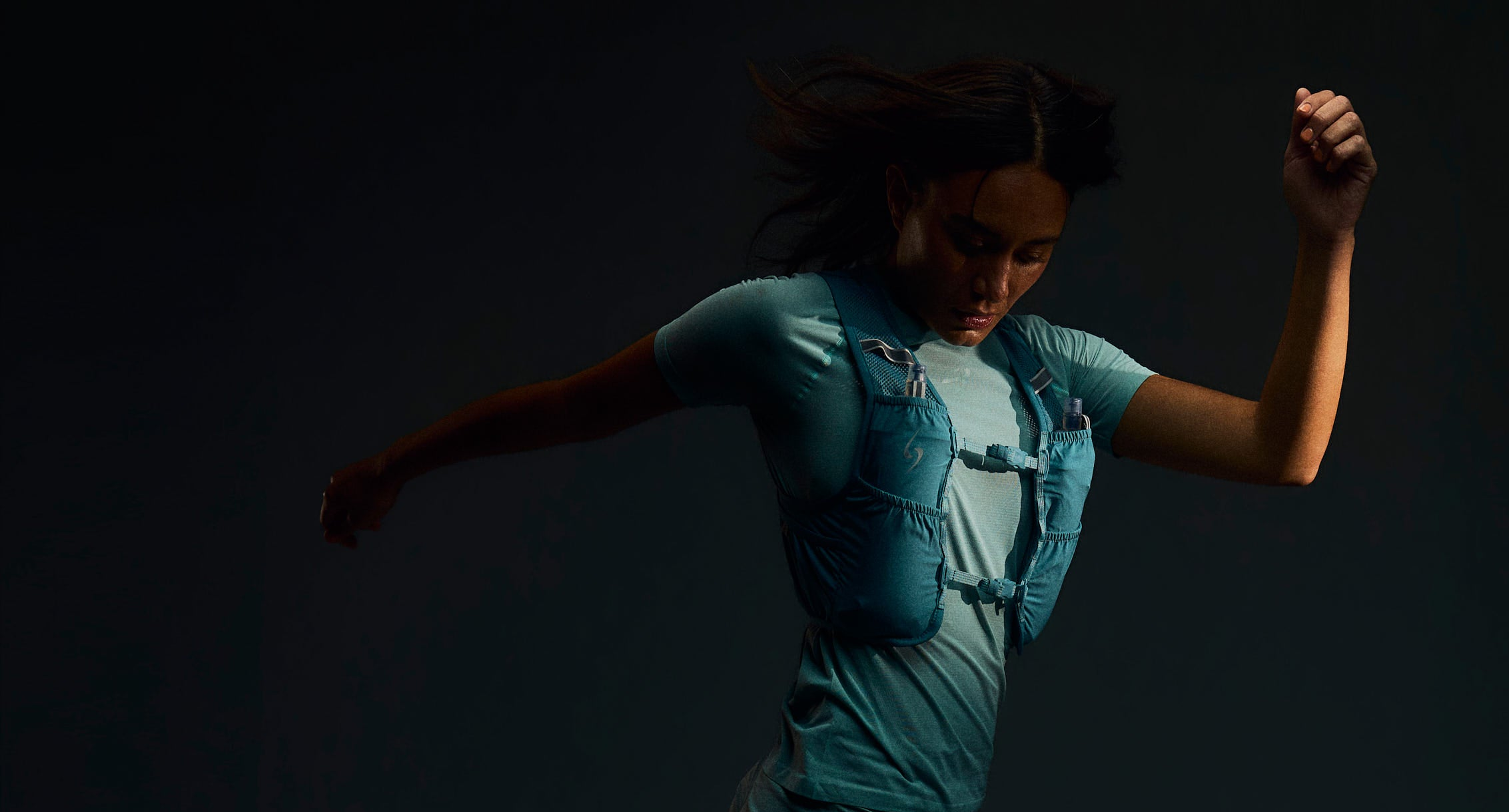A woman in a light blue athletic outfit and a light blue running vest is captured mid-stride against a very dark, near-black studio background.