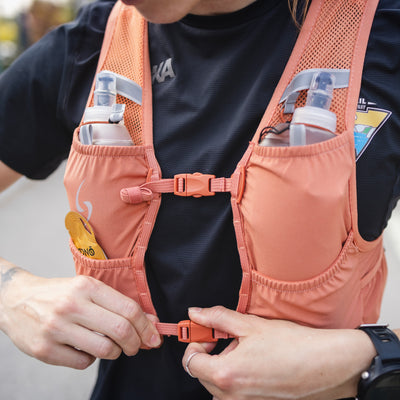 Person wearing a pink running vest with multiple pockets and water bottles, outdoors. #color_terra-cota