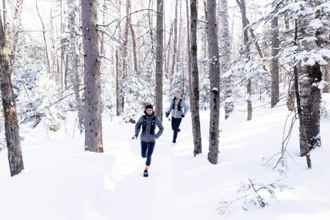 Femme et homme courant dans un sentier l'hiver, portant des vestes d'hydratation et tenant des bâtons de randonnée.