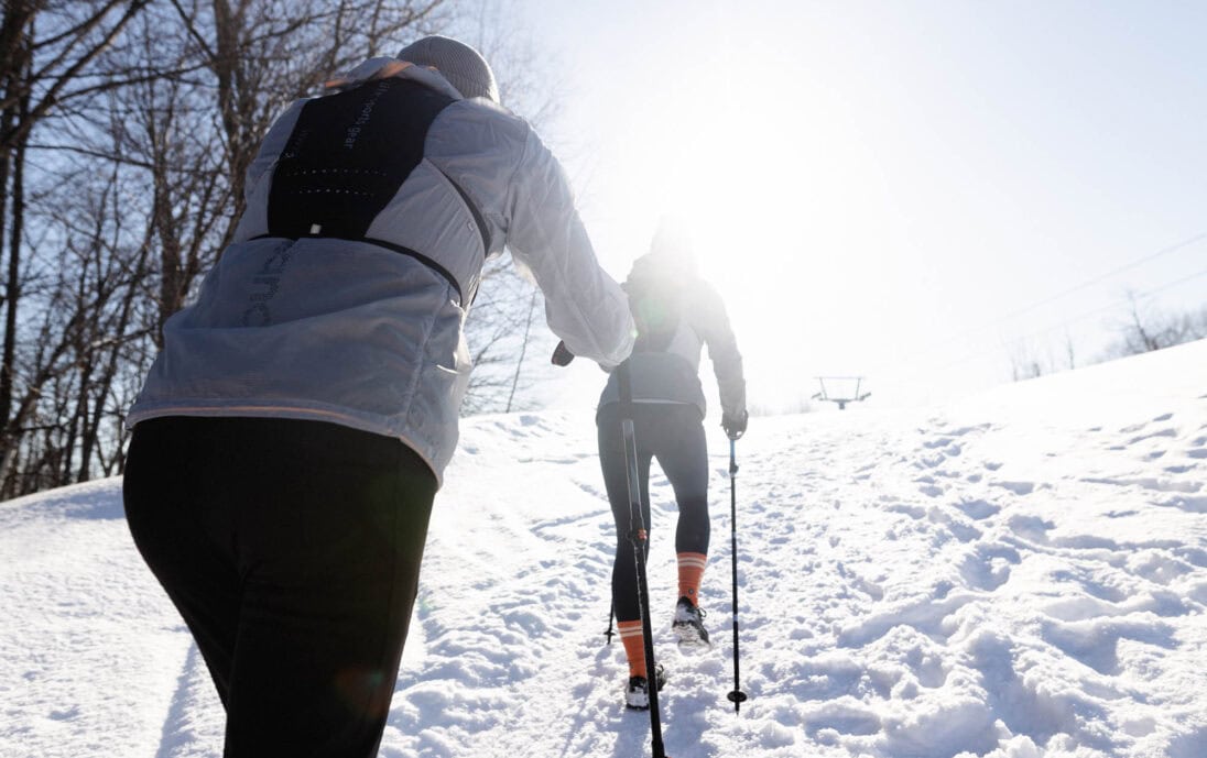 Deux randonneurs grimpent une montagne de ski enneigée à l'aide de bâtons de randonnée, par une journée ensoleillée.