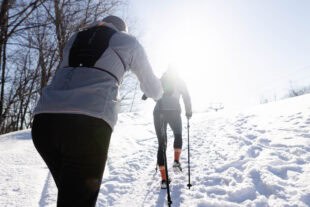 Deux randonneurs grimpent une montagne de ski enneigée à l'aide de bâtons de randonnée, par une journée ensoleillée.