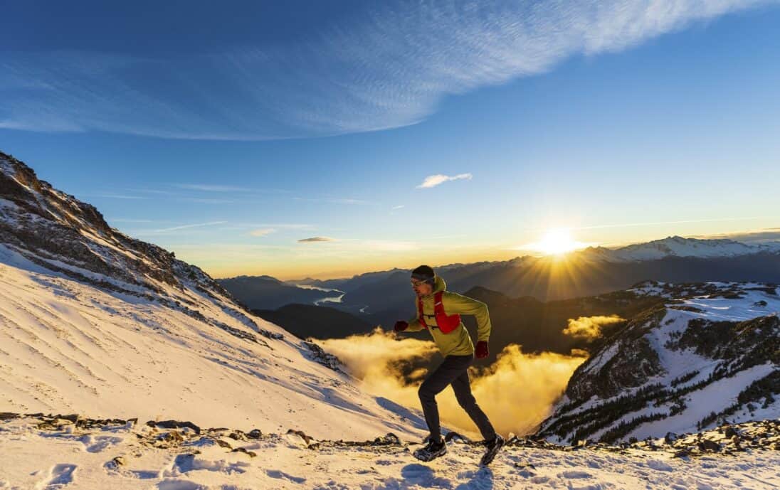 Jeune homme en randonnée alpine au coucher du soleil, portant crampons et veste d'hydratation, dans un magnifique paysage.