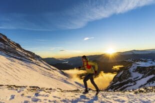 Jeune homme en randonnée alpine au coucher du soleil, portant crampons et veste d'hydratation, dans un magnifique paysage.